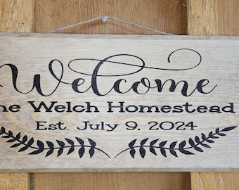 May include: A rectangular wooden sign with the word "Welcome" in elegant script. Below, it reads "The Welch Homestead Est. July 9, 2024" in a serif font, with a decorative leaf design. The sign is hanging.