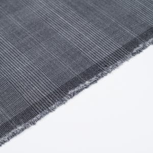 Può includere: Primo piano di una sezione di tessuto grigio con un motivo a quadri. Il tessuto ha un bordo sfilacciato ed è posto su uno sfondo bianco. La trama è visibile, mostrando la consistenza e i dettagli del materiale.