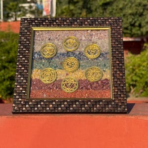 May include: A framed artwork featuring seven gold-colored chakra symbols set against a colorful, layered background of small stones. The frame is a dark brown, woven pattern. The artwork is displayed outdoors.