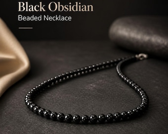 May include: A black obsidian beaded necklace featuring a single strand of polished, uniform black spherical beads. The text 'Black Obsidian Beaded Necklace' is displayed in the upper left corner of the image above a thin horizontal line.