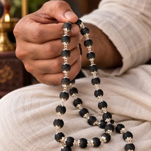 May include: A string of prayer beads featuring matte black spherical beads, each separated by intricate silver-toned metal caps. The beads are linked together in a continuous chain, showcasing a contrast between the dark, textured spheres and the bright, polished metallic accents throughout the design.