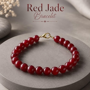 May include: A red jade bead bracelet featuring smooth, round, deep red stones strung together. The bracelet includes a gold-toned metal lobster claw clasp. The text "Red Jade Bracelet" is displayed in dark red lettering above the jewelry piece.
