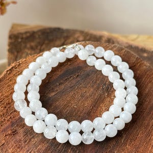 May include: A double strand necklace composed of white, round beads. The beads are strung together with a silver clasp. The necklace is displayed on a wooden surface, showcasing the jewelry's natural aesthetic.