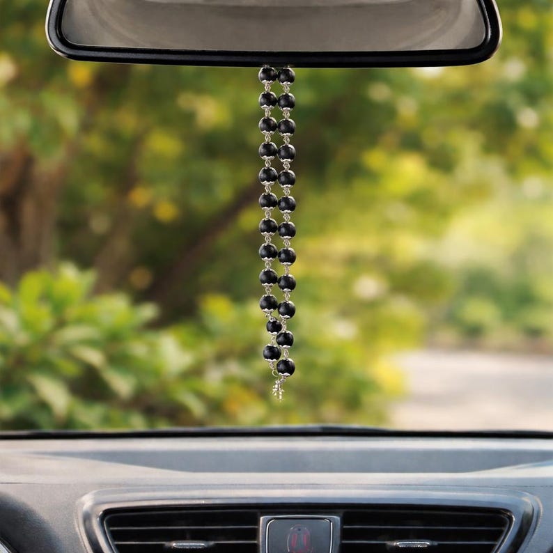 May include: A car rearview mirror hanging ornament featuring a double strand of matte black spherical beads separated by small silver-toned metal spacers, ending in a small silver-toned cross charm suspended from the vehicle mirror.
