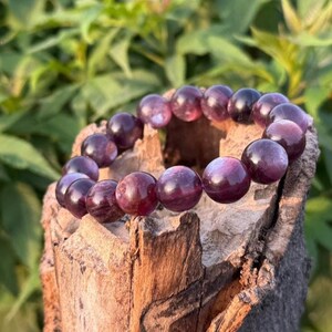 以下が含まれることがあります： 深い紫とバーガンディの色合いの、大きく丸く磨かれた宝石をあしらったビーズブレスレット。風化した木片の上にブレスレットが展示されており、背景はぼやけた緑色です。