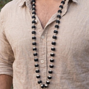 May include: A long necklace featuring a series of round, textured black beads separated by small, ornate silver-toned metal caps. The beads are strung together in a single strand that hangs down the centre of a light tan, button-down linen shirt.