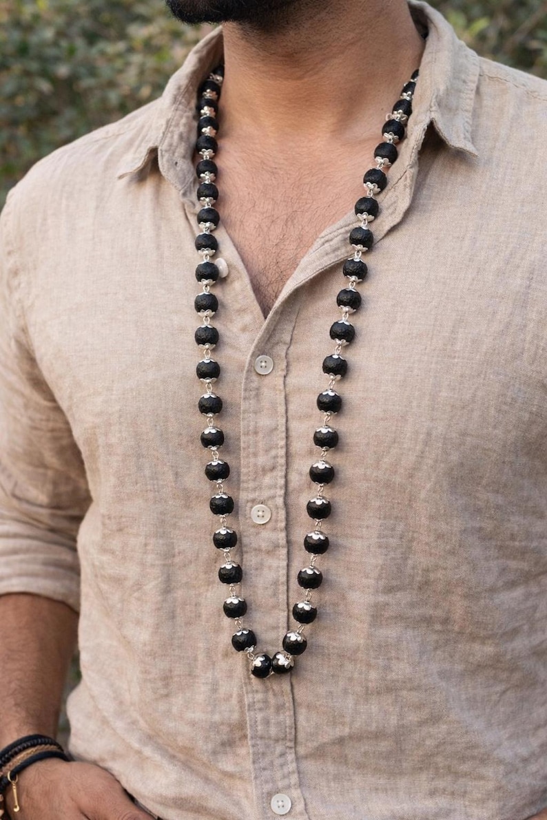 May include: A long necklace featuring a series of round, textured black beads separated by small, ornate silver-toned metal caps. The beads are strung together in a single strand that hangs down the centre of a light tan, button-down linen shirt.