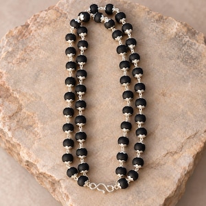 May include: A double-strand necklace featuring matte black spherical beads, each accented with small silver-toned metal caps at the top and bottom. The strands are connected by a silver-toned metal S-hook clasp at the base of the necklace.