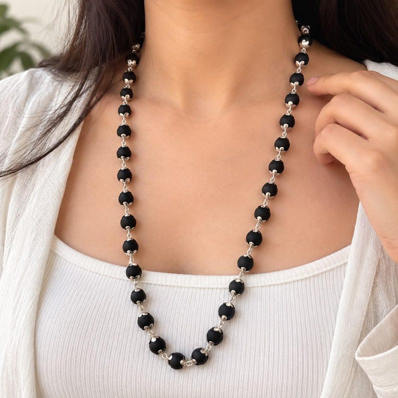 May include: A long necklace featuring a series of matte black spherical beads, each separated by small, intricate silver-toned metal caps and connected by thin silver-toned metal links, creating a uniform and repeating pattern around the neck.
