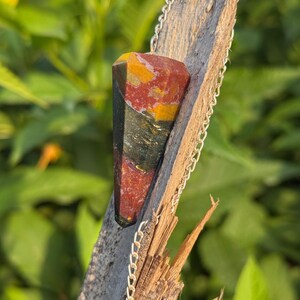 May include: A gemstone pendant with a silver chain. The pendant is a faceted, elongated shape with red, yellow, green, and black colors. The chain is attached to the top of the pendant and has a small, clear bead at the end.