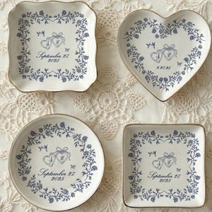 Può includere: Quattro piattini in ceramica di forma quadrata, a cuore e rotonda, caratterizzati da un bordo floreale blu e un disegno centrale di due cuori intrecciati con il testo September 27 2025. Ogni piattino ha un bordo dorato e una base color crema.