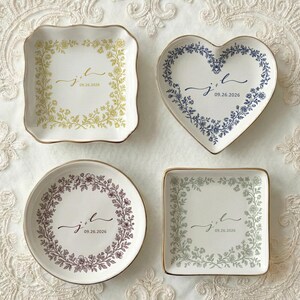 A top down view of four ceramic ring dishes with gold trim. One is heart shaped, one is round, one is scalloped and one is square shaped. On the ring dish is a floral a toile de jouy style border and cursive initials with a date under the initials.