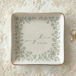 Könnte beinhalten: Ein quadratisches, cremefarbenes Keramiktablett mit goldenem Rand, verziert mit einem floralen Design und den Initialen "j + l" sowie dem Datum "09.26.2026". Zwei goldene Creolen mit Perltropfen liegen auf einer Spitzendecke.