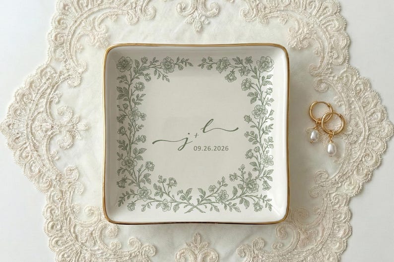 A top down view of a square ceramic ring dish with gold trim. On the ring dish is a floral a toile de jouy style border and cursive initials with a date under the initials.
