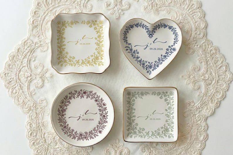 A top down view of four ceramic ring dishes with gold trim. One is heart shaped, one is round, one is scalloped and one is square shaped. On the ring dish is a floral a toile de jouy style border and cursive initials with a date under the initials.