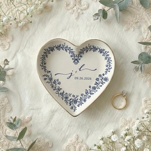 A top down view of a heart shaped ceramic ring dish with gold trim. On the ring dish is a floral a toile de jouy style border and cursive initials with a date under the initials.