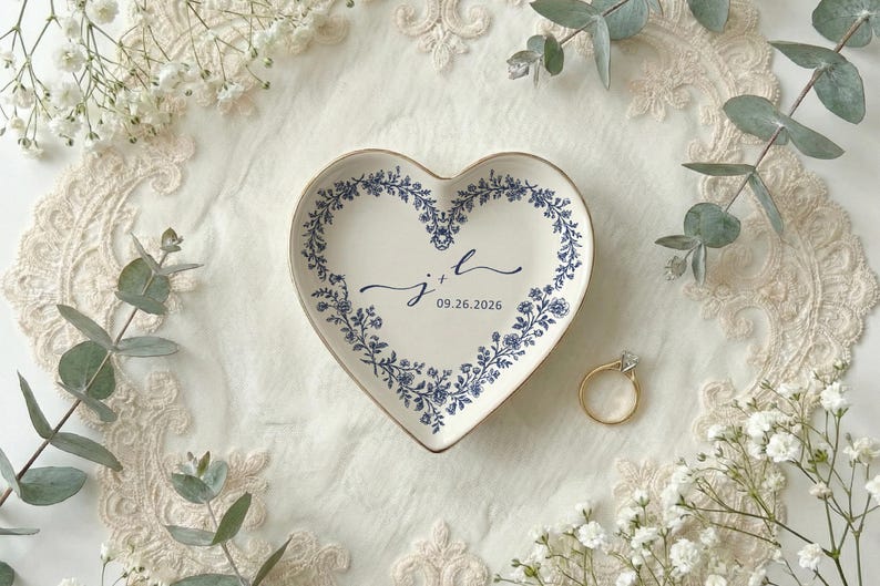 A top down view of a heart shaped ceramic ring dish with gold trim. On the ring dish is a floral a toile de jouy style border and cursive initials with a date under the initials.
