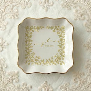 A top down view of a square shaped scalloped ceramic ring dish with gold trim. On the ring dish is a floral a toile de jouy style border and cursive initials with a date under the initials.