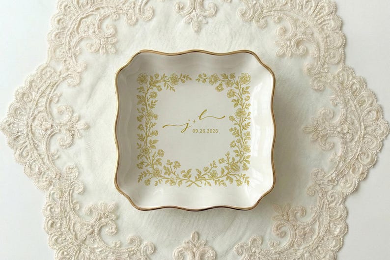 A top down view of a square shaped scalloped ceramic ring dish with gold trim. On the ring dish is a floral a toile de jouy style border and cursive initials with a date under the initials.