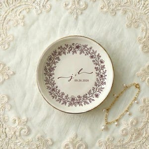 A top down view of a round ceramic ring dish with gold trim. On the ring dish is a floral a toile de jouy style border and cursive initials with a date under the initials.