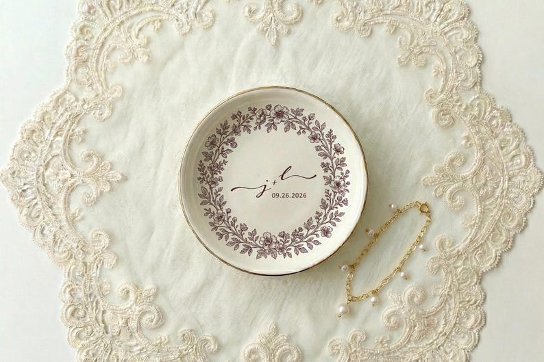 A top down view of a round ceramic ring dish with gold trim. On the ring dish is a floral a toile de jouy style border and cursive initials with a date under the initials.