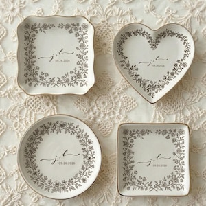 May include: Four decorative ceramic dishes in various shapes: square, heart, and round. Each dish is white with a floral border and personalized with initials and a date in black script. The edges have a gold trim.