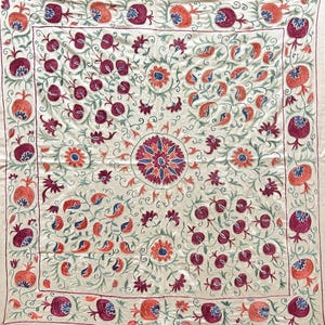 Könnte beinhalten: Cremefarbener Stoff mit aufwendiger Stickerei. Das Design zeigt Granatäpfel, Blumen und wirbelnde Ranken in Rot-, Blau- und Grüntönen. Ein zentrales Blumenmedaillon ist von sich wiederholenden Mustern umgeben.