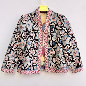 Peut inclure: Une veste à manches longues avec une base noire et un motif floral géométrique répétitif dans des tons de bleu, pêche et blanc. Le vêtement présente une bordure contrastée à motifs rouges et blancs sur l'ouverture avant et les poignets, avec une doublure jaune.