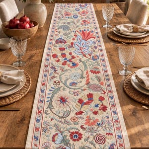 May include: A long rectangular table runner featuring an intricate embroidered floral pattern in shades of red, blue, and soft green on a cream-colored fabric base, finished with a thin red border along the length of the runner.