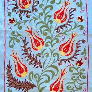 May include: A light blue textile with an embroidered floral design. The design features red tulips with yellow centers, green leaves, and dark brown stems. The edges are trimmed with a red border.