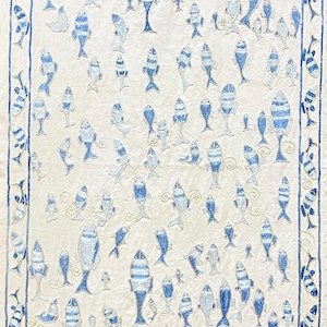 May include: A rectangular off-white textile featuring an embroidered pattern of numerous fish in varying shades of blue. The fish are arranged in a scattered formation across the centre, enclosed by a decorative border of fish motifs.