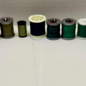 May include: A collection of various spools of thread in a variety of colors, including black, green, and white. The spools are arranged in a curved line, with some featuring labels. The thread spools are for sewing and crafting.