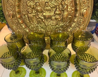 May include: A collection of green-tinted glassware, including goblets and tumblers, arranged in front of a large, ornate gold-colored metal platter. The goblets have diamond-patterned bases and stems. The platter features a detailed relief scene.