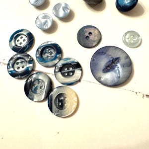 May include: A collection of various buttons in shades of blue, gray, and white. The buttons vary in size and design, including round, oval, and flat styles. Some buttons have four holes, while others have two. The buttons are arranged on a light-colored surface.