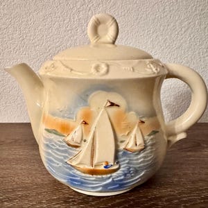 May include: Cream-colored ceramic teapot with a lid and handle. The teapot features a nautical design with sailboats on a blue and white ocean scene. The lid has a loop handle, and the spout is on the left side.
