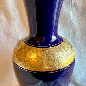 May include: A tall, elegant vase with a deep blue glaze and gold accents. The vase features a wide, gold band with a detailed floral pattern around the middle. The rim and base are also trimmed in gold, creating a luxurious look.