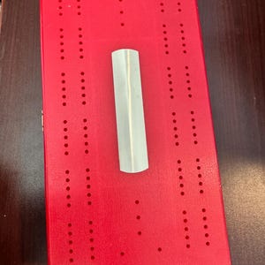 Puede incluir: Un tablero de cribbage rojo con múltiples filas de agujeros para insertar clavijas. Una clavija de color plateado se coloca en el centro del tablero. El tablero es rectangular y de madera. Este juego clásico es perfecto para las noches de juegos familiares.