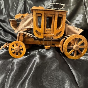 May include: A detailed wooden stagecoach model with a tan canvas top and wooden wheels. The stagecoach features intricate details, including chains and a driver's seat. The model is set against a dark, textured background.