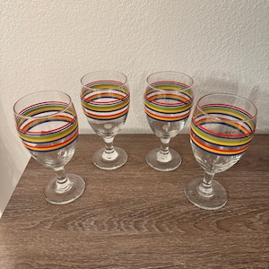May include: Four clear wine glasses with colorful horizontal stripes in red, yellow, blue, and green. The glasses have a classic stemmed design and are arranged on a wooden surface.