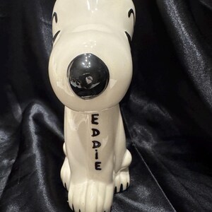 May include: A ceramic figurine of a seated dog with a glossy white finish. The dog has a large black nose, black eyes, and black markings. The name "EDDIE" is written vertically in black letters on the dog's chest.