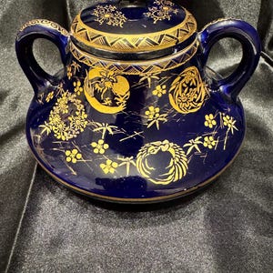May include: A dark blue ceramic sugar bowl with a lid and two handles. The bowl and lid are decorated with gold floral and geometric patterns. The lid has a small gold knob.