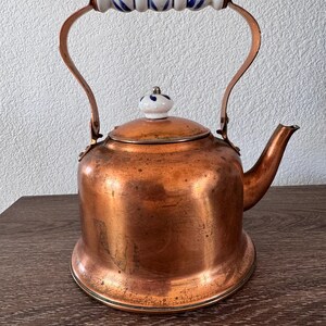 May include: A copper tea kettle with a wide base and a curved spout. The kettle features a white ceramic handle and a matching lid knob, both decorated with a blue floral pattern. The copper body shows a natural patina with visible surface wear.