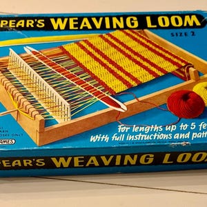 Op de afbeelding: Een vintage Spear's Weaving Loom maat 2 doos. De blauwe doos heeft een houten weefgetouw met rood en geel geweven stof, garen en de tekst "SPEAR'S WEAVING LOOM". Inclusief instructies en een patroonboek.