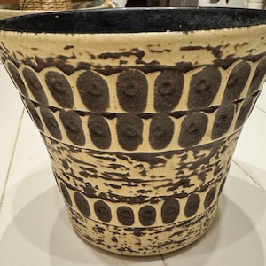 May include: A ceramic planter with a wide, slightly flared shape. The exterior features a cream-colored base with dark brown, oval-shaped patterns and textured accents. The rim is dark blue, creating a contrast. This decorative planter is suitable for indoor or outdoor use.