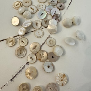 Può includere: Una collezione di bottoni vintage di varie dimensioni e design. I bottoni sono principalmente bianco sporco e color crema, alcuni con superfici iridescenti in madreperla. Alcuni bottoni hanno due o quattro fori, altri motivi decorativi.