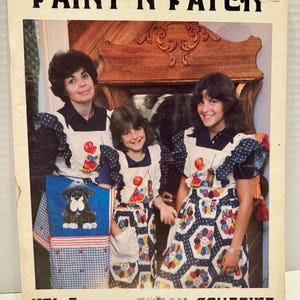 May include: A vintage book cover titled "Paint N' Patch" featuring three people wearing aprons with colorful patterns. The cover includes the text "VOL. 7" and "SUSAN SCHEEWE". The aprons have a dog design and fruit designs.