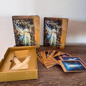 Peut inclure: Le jeu de cartes The Faeries Oracle de Brian Froud et Jessica Macbeth, comprenant une boîte de rangement dorée, un livre relié et une collection de cartes oracle illustrées aux tons bleus et dorés, disposées en éventail.