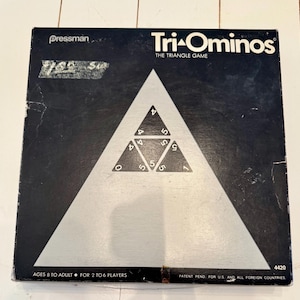 May include: A vintage Tri-Ominos board game box. The box is black with a large white triangle in the center. Inside the triangle are smaller triangles with numbers. The game is for ages 8 to adult and for 2 to 6 players.