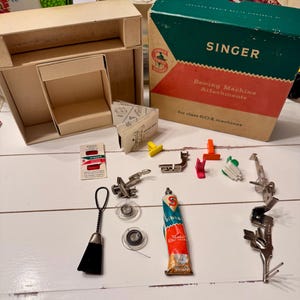 May include: A collection of vintage Singer sewing machine attachments and accessories, including a box, instruction booklet, bobbins, and a tube of oil. The box is labeled "Singer Sewing Machine Attachments". Various colored presser feet are also visible.