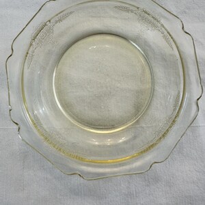 Può includere: Un piatto di vetro trasparente e vintage con bordo smerlato e un motivo decorativo inciso. Il piatto ha una tinta leggermente gialla e un centro circolare. Il design inciso presenta elementi floreali e geometrici.
